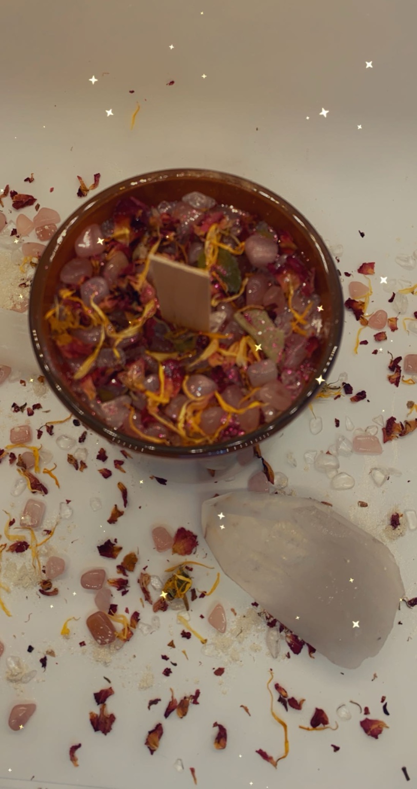 Overhead view of a raw intention candle topped with crystals and florals scattered across a white surface.
